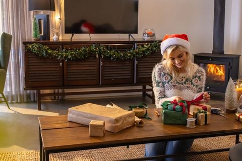 Woman wearing santa hat wrapping gifts in cozy living room with fireplace