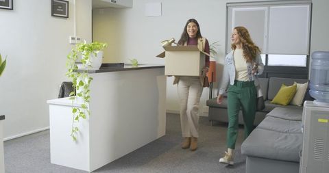 Female Coworkers Carrying Cardboard Box Through Modern Office Reception Smiling and Talking