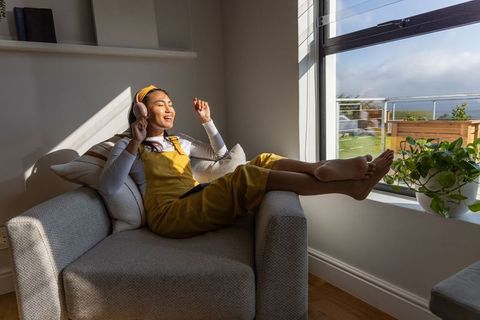 Asian woman relaxing in sunlit armchair wearing yellow overalls listening with headphones