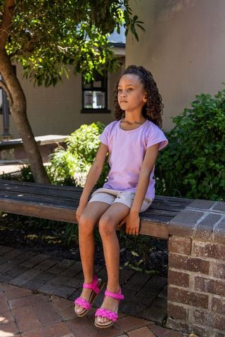 African american school-age girl sitting on wooden bench in sunlit courtyard wearing pink sandals