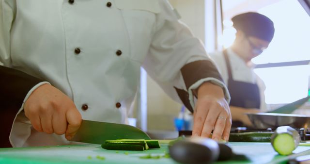 Chefs wearing traditional chef coats cutting fresh vegetables on a green chopping board in a professional kitchen. This image showcases culinary skills, teamwork, and food preparation processes. Perfect for use in articles about gastronomy, culinary arts, restaurant business, and cooking classes.