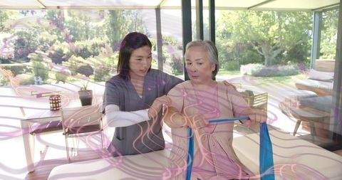 Therapist assisting asian senior exercising with resistance band in sunlit rehab room