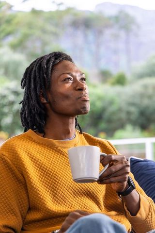Contemplative african american man sipping coffee on porch in mustard sweater and smartwatch