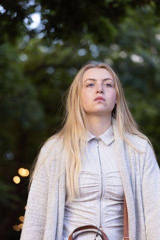 Contemplative blonde woman standing in garden wearing cardigan and leather bag with bokeh lights