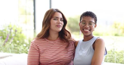 Smiling multiracial women sitting together on sunlit sofa sharing warm friendship moment