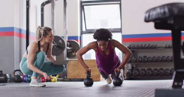 Image of diverse female fitness trainer instructing woman doing push ups on kettlebells at gym. Exercise, fitness and healthy lifestyle.