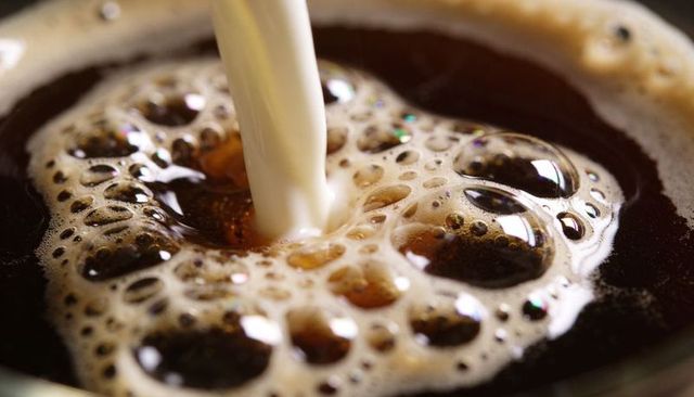 Pouring milk creating swirling foam and glossy bubbles in dark coffee cup macro closeup
