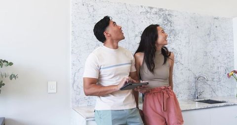 Diverse couple exploring modern kitchen holding tablet and smiling by marble backsplash