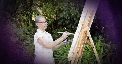 Senior chinese woman painting at garden easel, creating art in lush greenery