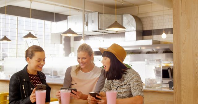 Three young women gather around a table in a contemporary café, engaging with their smartphones while enjoying pink milkshakes. Ideal for illustrating themes of friendship, modern social interactions, and the casual leisure culture. Perfect for marketing materials, blogs, and advertisements focused on youth lifestyle, technology, and dining out.