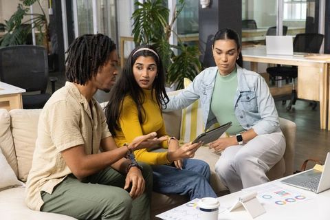 Multiracial team collaborating over tablet and charts on office sofa in coworking lounge