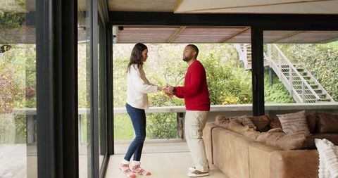 Couple holding hands and dancing near floor-to-ceiling glass doors in modern living room