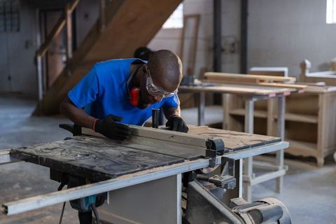 Craftsman cutting wooden plank on table saw in busy carpentry workshop