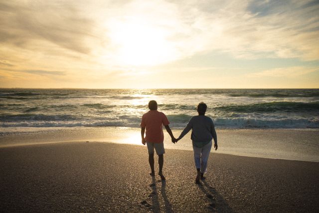 Senior couple holding hands walking towards the shore at sunset. Ideal for themes of love, togetherness, retirement, and peaceful lifestyles. Perfect for use in advertisements, brochures, or articles related to senior living, romantic getaways, or healthy aging.