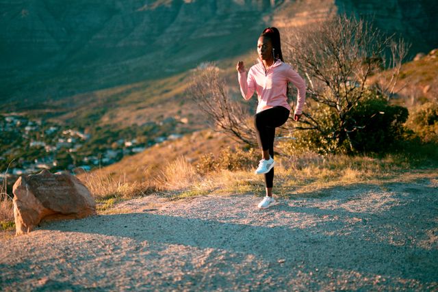 African American woman wearing sportswear and wireless earphones, exercising in a scenic countryside setting. Ideal for promoting healthy lifestyles, outdoor fitness, and wellness activities. Suitable for use in fitness blogs, health magazines, and advertisements for sportswear or fitness equipment.