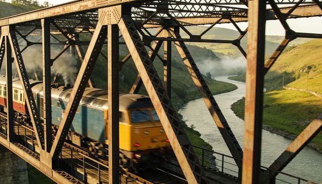 Yellow blue diesel locomotive crossing riveted steel truss bridge over winding misty river at sunris