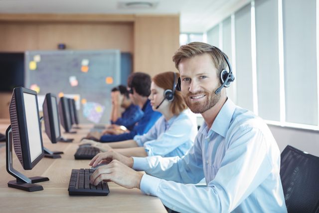 Businessman wearing headset smiling while working at desk in call center. Ideal for illustrating customer service, teamwork, and professional office environments. Useful for corporate websites, business presentations, and marketing materials related to customer support and communication.