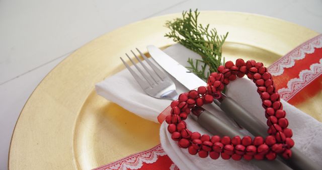 A festive table setting features a golden charger plate and silverware wrapped in a napkin, adorned with a red heart-shaped bead decoration, with copy space. It suggests a romantic dinner or a special celebration such as Valentine's Day or an anniversary.