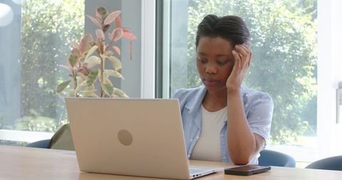 Young black woman working on laptop at bright home office with plant and smartphone