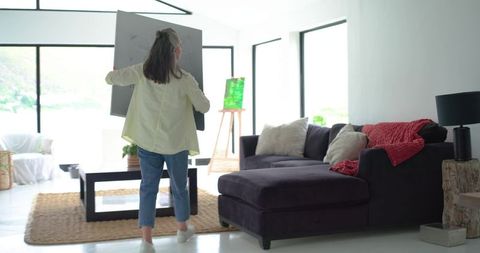 Mature woman carrying large canvas into bright modern living room for artwork display