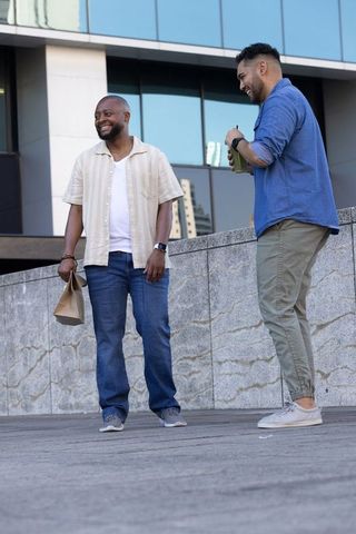 Two diverse friends chatting on urban plaza holding takeout bag and reusable cup