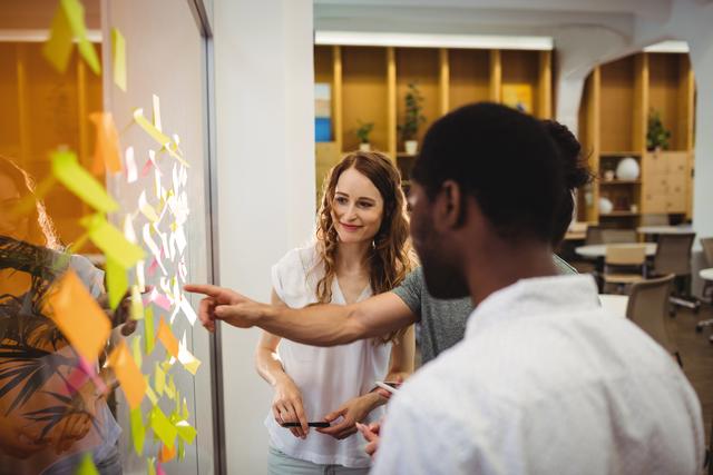 Diverse group of business professionals collaborating and brainstorming with sticky notes on glass wall in modern office. Ideal for illustrating teamwork, creative processes, project management, and business strategy in corporate environments.