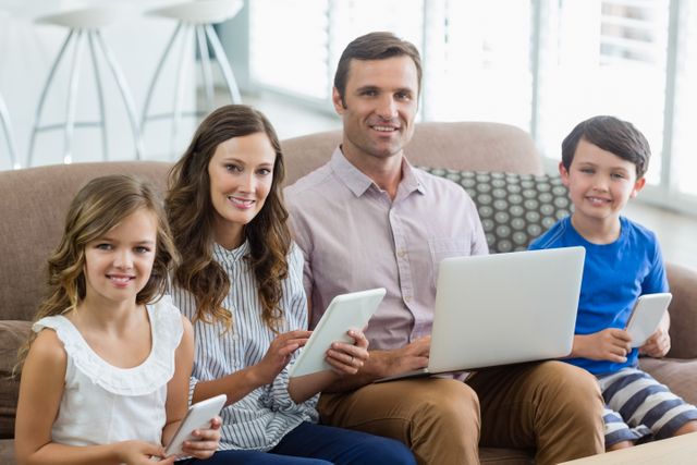 Group portrait of a happy family sitting on a sofa using digital devices in a bright and cozy living room. The image highlights family bonding time while engaging with technology. Perfect for illustrating concepts related to modern family life, technology use, and home lifestyle.
