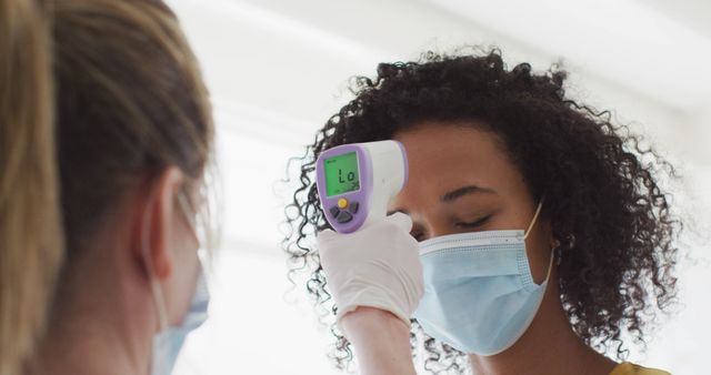 Image depicts a healthcare worker wearing protective gloves checking a patient's temperature using an infrared thermometer. Patient, with curly hair, is wearing a face mask. Perfect for content related to health screenings, pandemic measures, medical facilities, or promoting health and safety protocols during the COVID-19 pandemic.