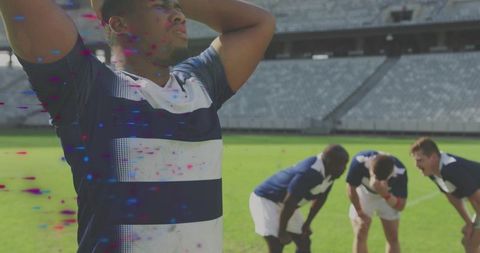 Rugby player holding head with exhausted teammates bending on grass in empty stadium