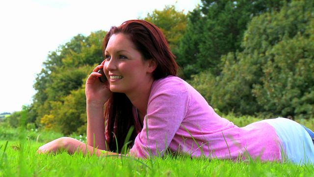 Woman engages in a phone conversation while lying on grass. Ideal for themes of relaxation, leisure activities, or technological communication in nature. Useful for promoting lifestyle content, advertisements for phone services, or articles focusing on outdoor experiences.