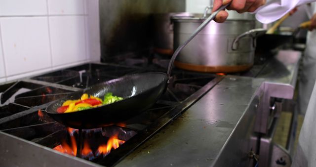 Chef cooking freshly cut vegetables in frying pan over open flame in commercial kitchen. Useful for illustrating culinary arts, restaurant promotion, professional cooking services, or culinary school training materials.