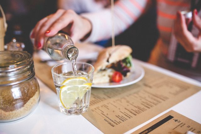 Person pouring water into glass with lemon slice on table, next to plate with sandwich and vegetables and a restaurant menu. Ideal for illustrating themes of dining, refreshment, casual meals, or healthy eating.