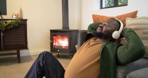 African american man relaxing with headphones by glowing wood stove in cozy living room