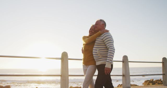 Senior couple walking arm in arm by the seaside during a beautiful sunset. Ideal for use in advertisements promoting elderly lifestyle, retirement services, or travel destinations aimed at older adults. Can also be used in inspirational materials focused on love, companionship, and active aging.
