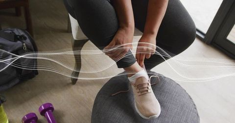 Woman tying sneaker on ottoman before home workout with dumbbells and gym bag