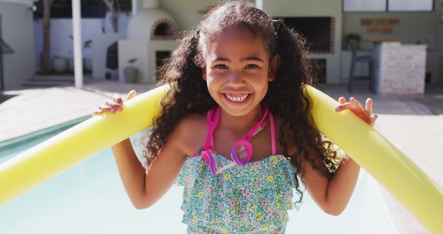 A young girl with curly hair smiles as she holds onto a yellow pool noodle by the poolside. She wears a floral swimsuit and looks ready for summer fun. This image is perfect for promotions or advertisements related to children's summer activities, swim gear, vacation destinations, and family leisure.