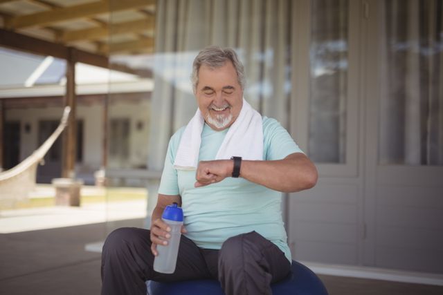 Senior man sitting on veranda after workout, checking time on wristwatch. He is holding a water bottle and has a towel around his neck, indicating a recent exercise session. This image is perfect for promoting active lifestyles, fitness routines for older adults, health and wellness campaigns, and outdoor relaxation.