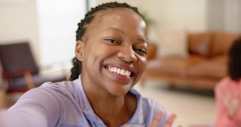 Smiling young woman taking selfie and waving in cozy living room wearing purple top
