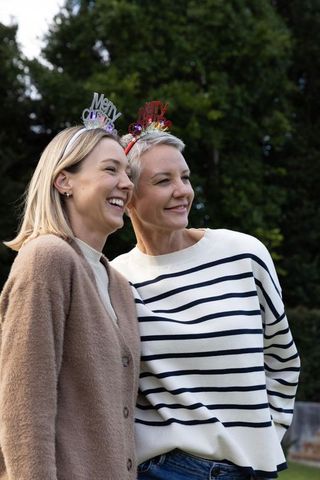 Two women laughing and leaning together wearing festive 'merry' headbands in garden