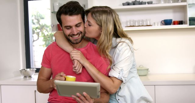 Young couple embracing in a bright kitchen while man is holding a tablet and a cup of coffee. Woman gives a kiss on the cheek exhibiting affection and love. Suitable for use in advertisements for home products, technology, coffee brands, or lifestyle blogs promoting love and togetherness.