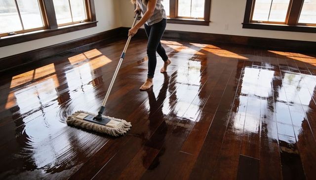 Barefoot woman mopping glossy hardwood floor in sunlit empty room with reflections