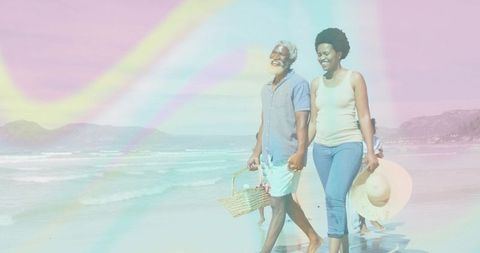African american couple walking along pastel beach holding picnic basket and hat, smiling