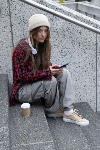 Young woman sitting on urban steps using smartphone with headphones and coffee cup