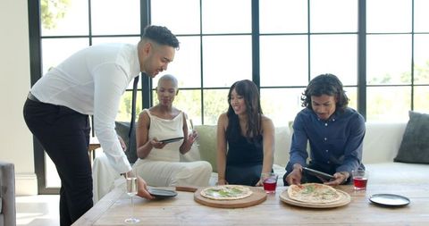 Man placing plate as friends share pizza in sunlit modern minimalist living room