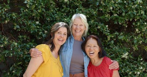 Three women friends smiling and embracing outdoors in front of flowering vine wall