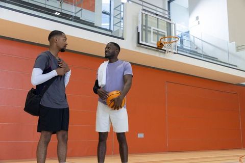 Two african american men talking and holding basketball on indoor gym court with hoop