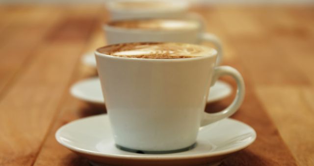 Cups of coffee with frothy tops arranged in a line on a wooden table, creating a warm and inviting atmosphere. Suitable for use in cafe menus, food and drink blogs, and advertisements for coffee products.