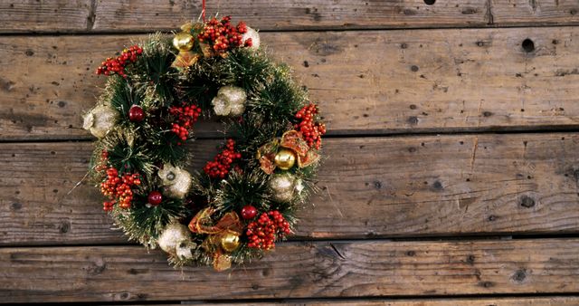 A festive Christmas wreath adorned with gold ornaments and red berries hangs on a rustic wooden wall, with copy space. Its traditional holiday decor adds a warm, seasonal touch to the setting.