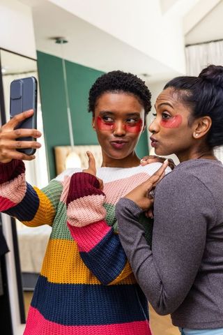Diverse friends taking mirror selfie wearing hydrogel under-eye patches and colorful knit sweater