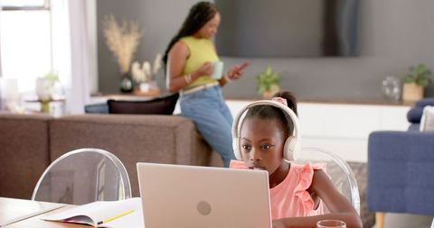 African daughter studying on laptop with white headphones while mother supervising at home